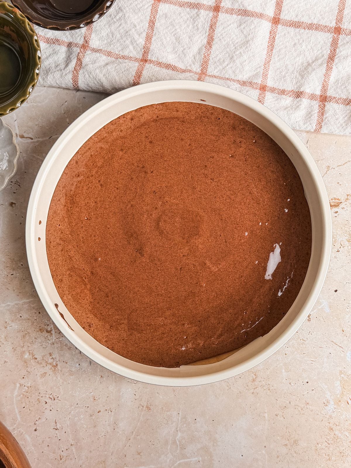 A round baking pan filled with smooth genoise chocolate cake batter sits on a light marble countertop next to a beige and orange plaid kitchen towel.