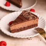 A slice of easy chocolate mousse cake with chocolate shavings sits on a white plate, accompanied by fresh raspberries and a gold fork. Another plate with cake is visible in the background.