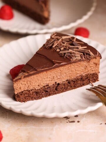 A slice of easy chocolate mousse cake with chocolate shavings sits on a white plate, accompanied by fresh raspberries and a gold fork. Another plate with cake is visible in the background.
