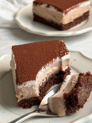 Slice of chocolate mousse cake on a small plate with a layer of cake, thick layer of mousse, whipped cream and cocoa powder