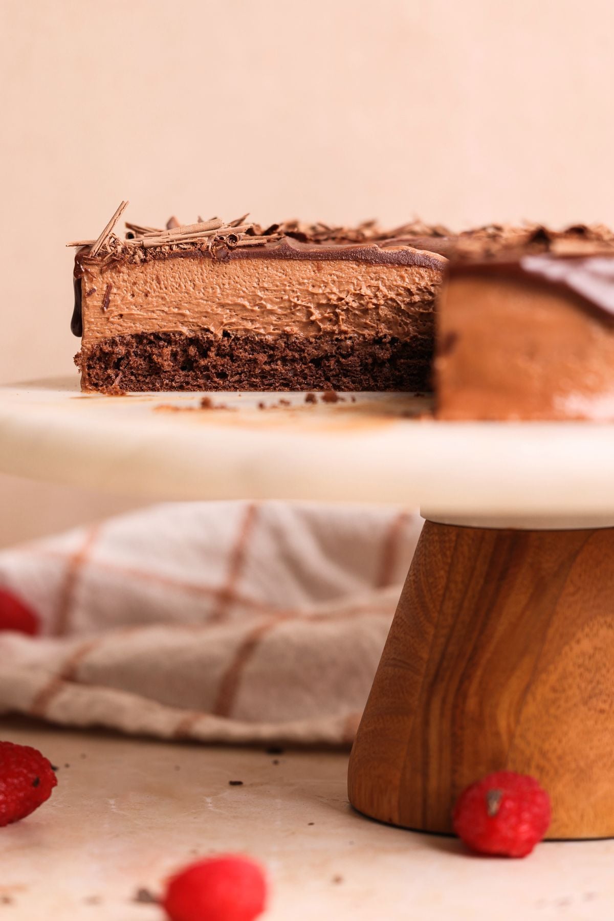 A chocolate mousse cake with a chocolate genoise base, partly sliced to reveal its creamy texture and rich ganache, sits on a cake stand. A beige cloth and scattered raspberries are visible in the background.