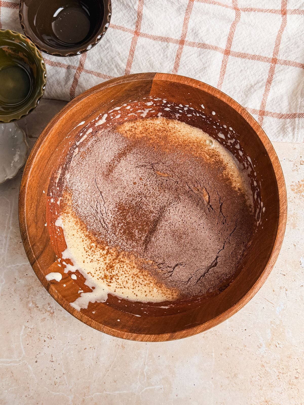 A wooden bowl containing a mixture of flour, cocoa powder, and liquid ingredients sits on a light countertop, with empty bowls and a checkered cloth nearby.