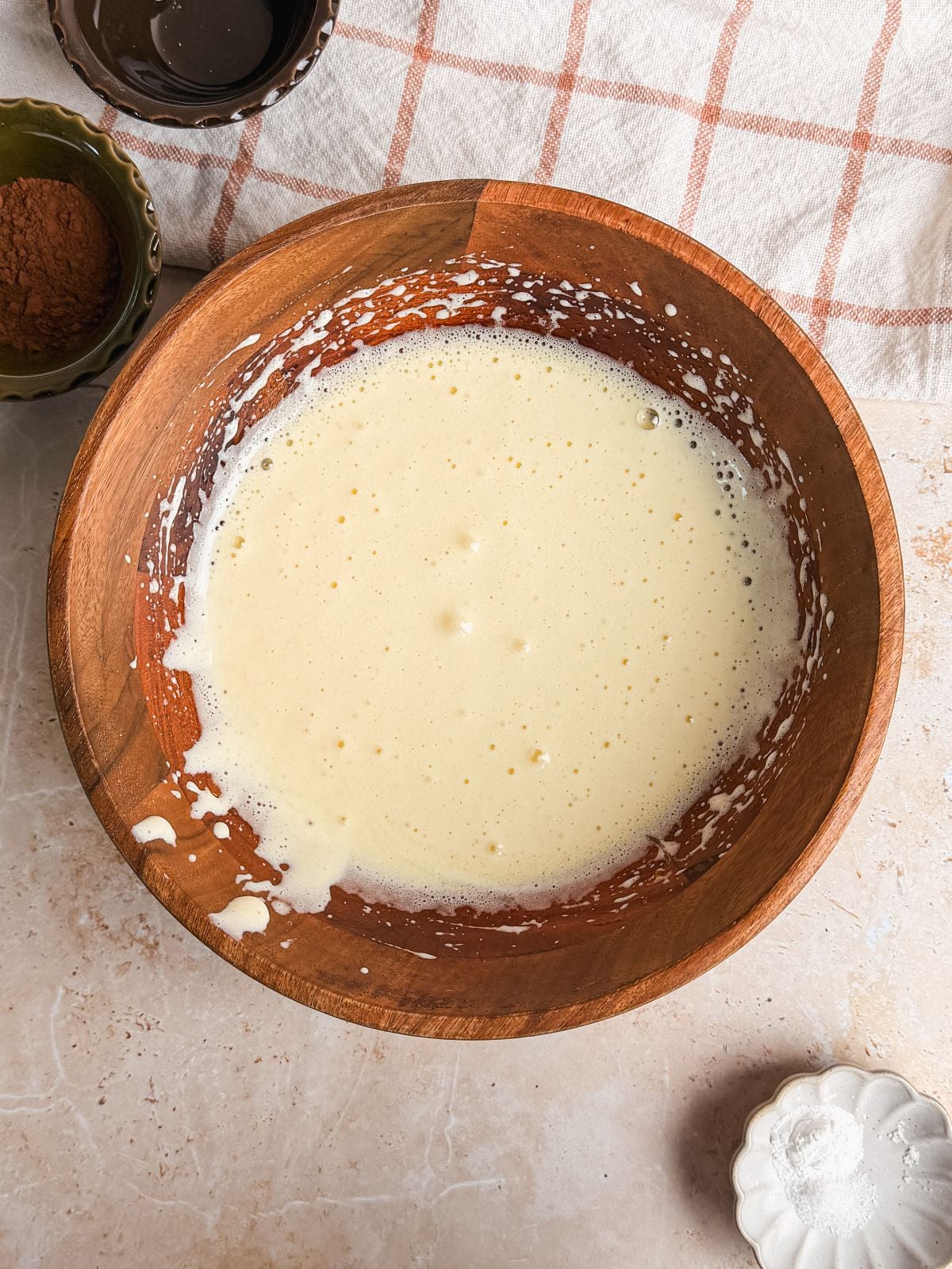 A wooden bowl with a creamy, pale mixture of egg and sugar sits on a light countertop. A beige plaid dish towel is partially visible in the background.