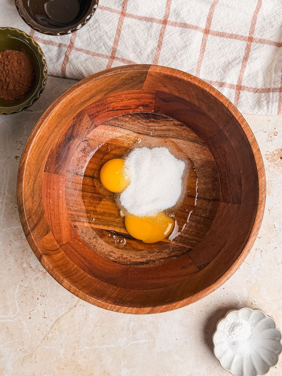 A wooden bowl with two raw eggs and a mound of sugar on a beige countertop, surrounded by small bowls of cocoa powder and salt, and a cloth with a checkered pattern.