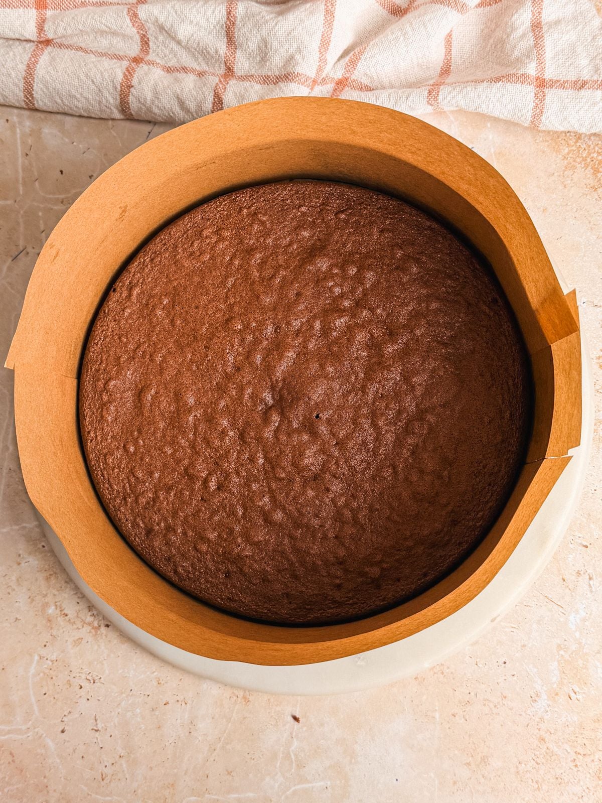 A freshly baked round chocolate genoise cake in a pan lined with brown parchment paper, placed on a light-colored countertop with a beige and white checkered cloth in the background.