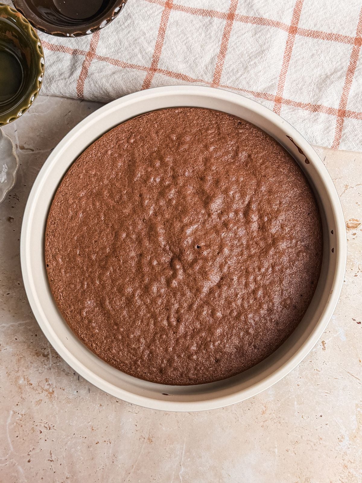 A round genoise chocolate cake in a white baking pan sits on a light countertop next to a beige and orange plaid kitchen towel.