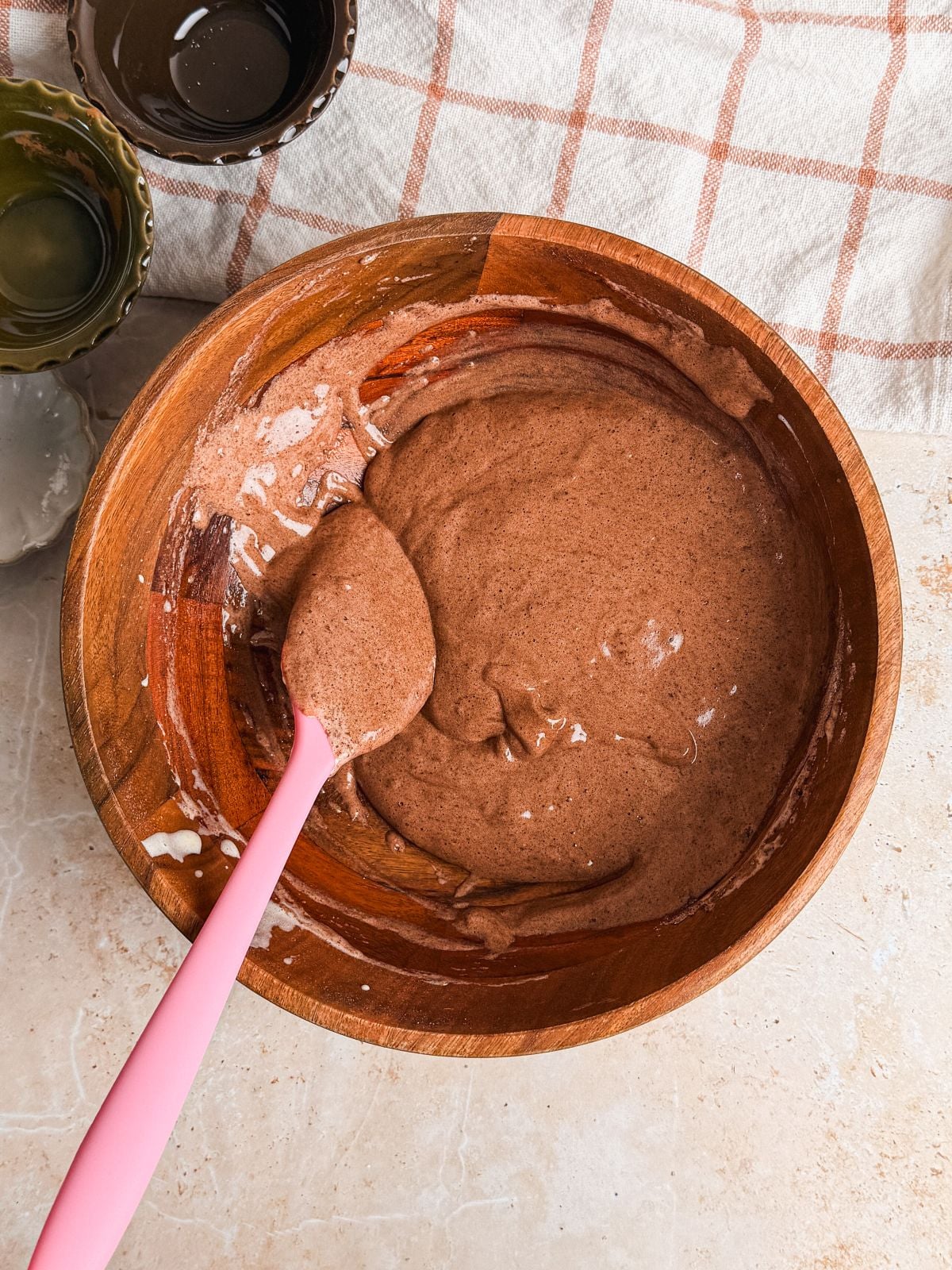 A wooden bowl filled with chocolate genoise batter and a pink spatula resting inside. The bowl sits on a light countertop with a beige and white checkered cloth nearby, along with a few small bowls.
