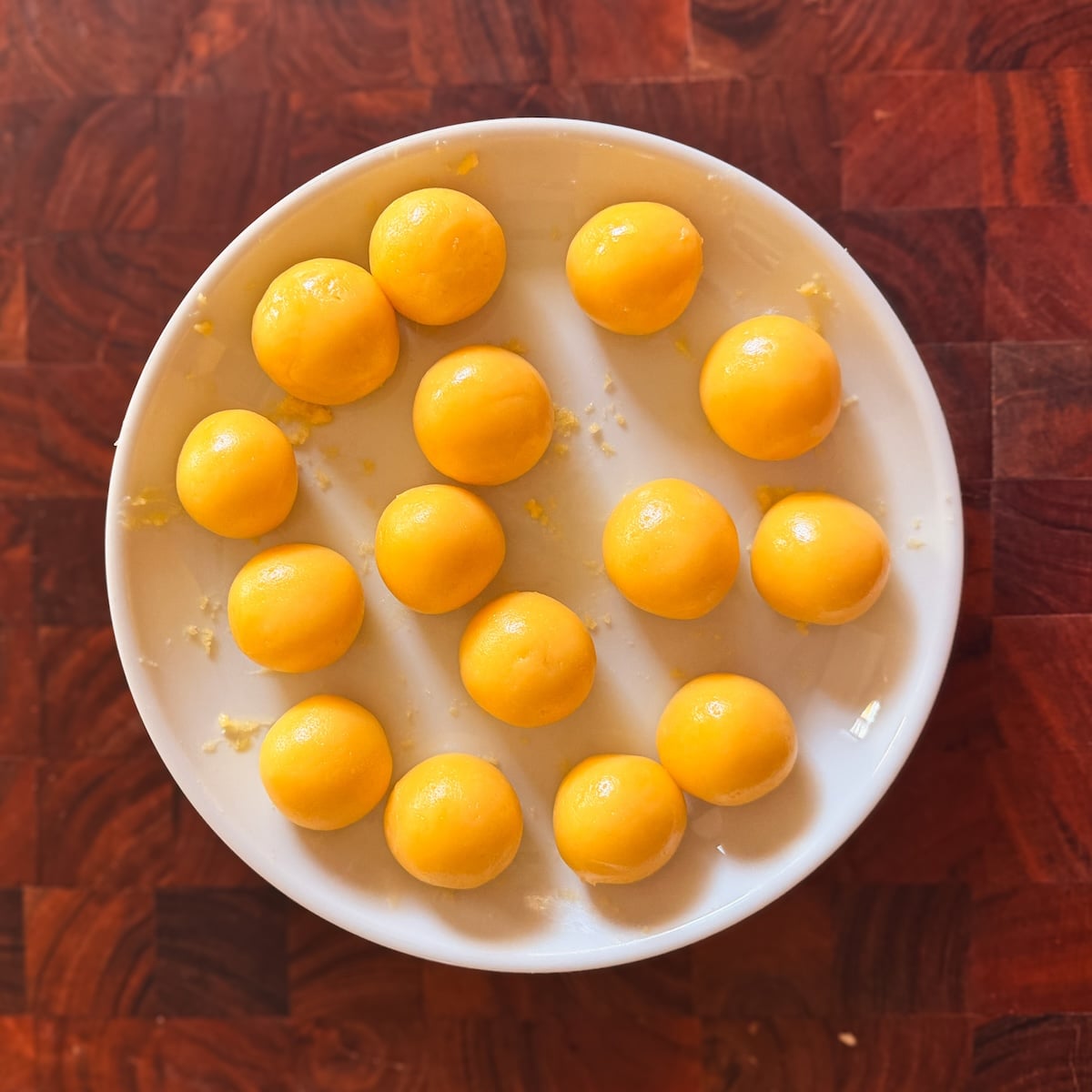 A white plate holds sixteen round, yellow balls of easy homemade gulab jamun arranged in a loose grid. The plate sits on a reddish-brown wooden surface with a geometric pattern.