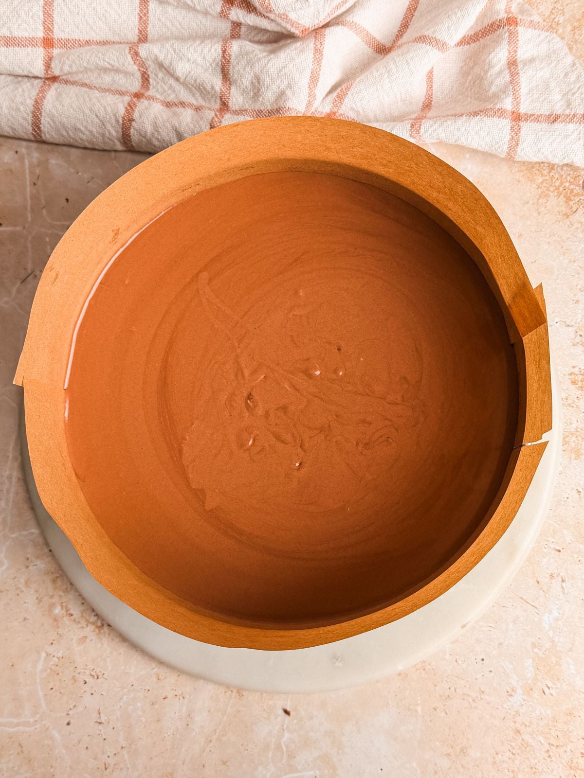 A round genoise chocolate cake in a white baking pan sits on a light countertop next to a beige and orange plaid kitchen towel.