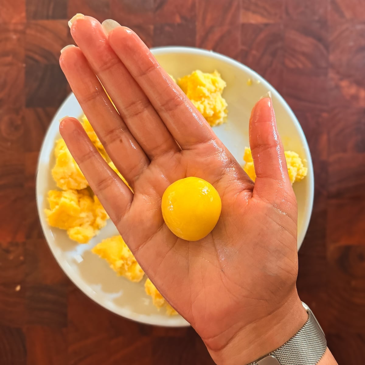 A hand holding a small, smooth yellow ball of dough over a plate with several pieces of similar yellow dough on a wooden surface, ready for easy homemade gulab jamun.