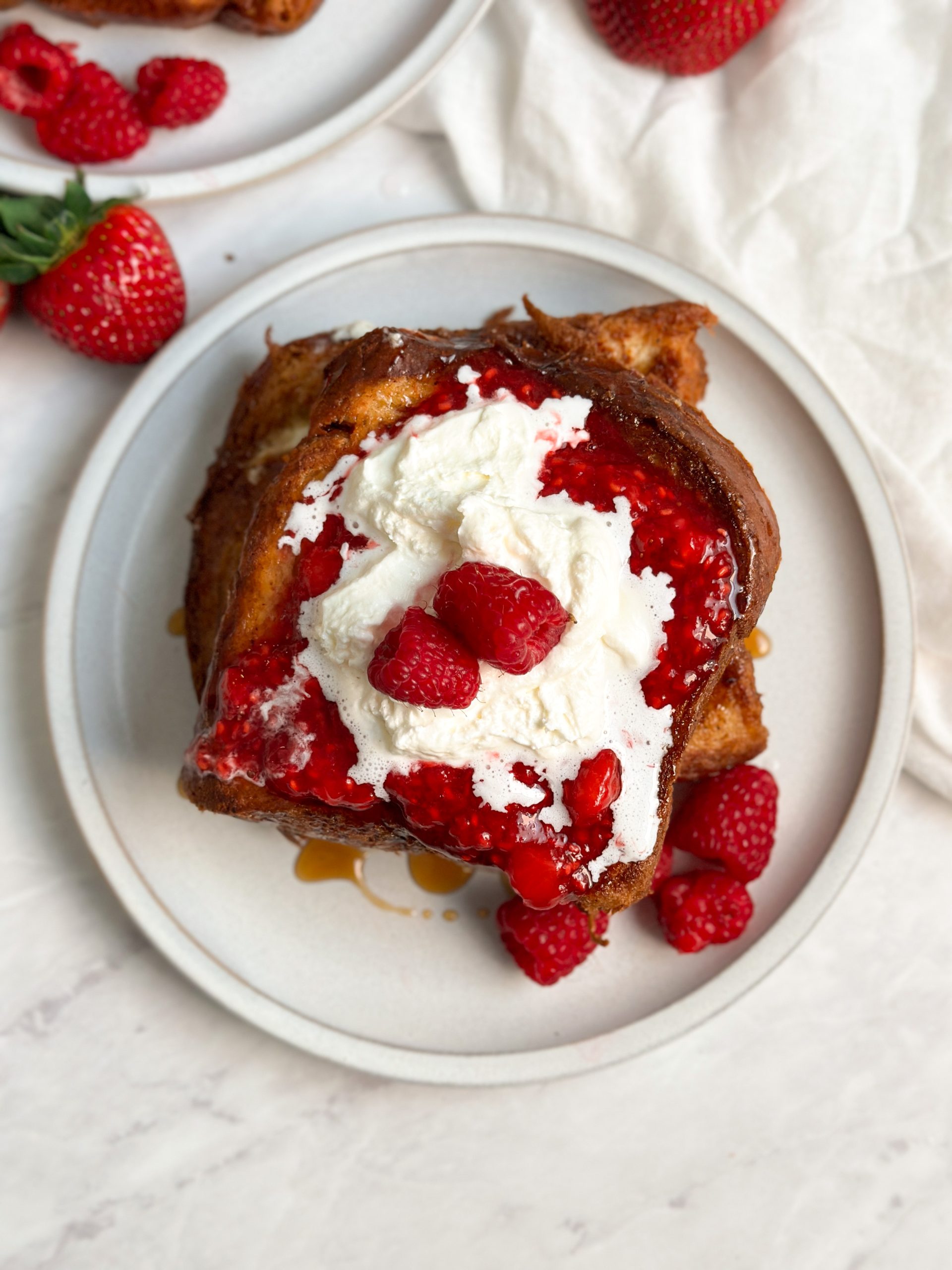 2 slices of cheesecake french toast on a plate with a berry compote, whipped cream and fresh raspberries from the top