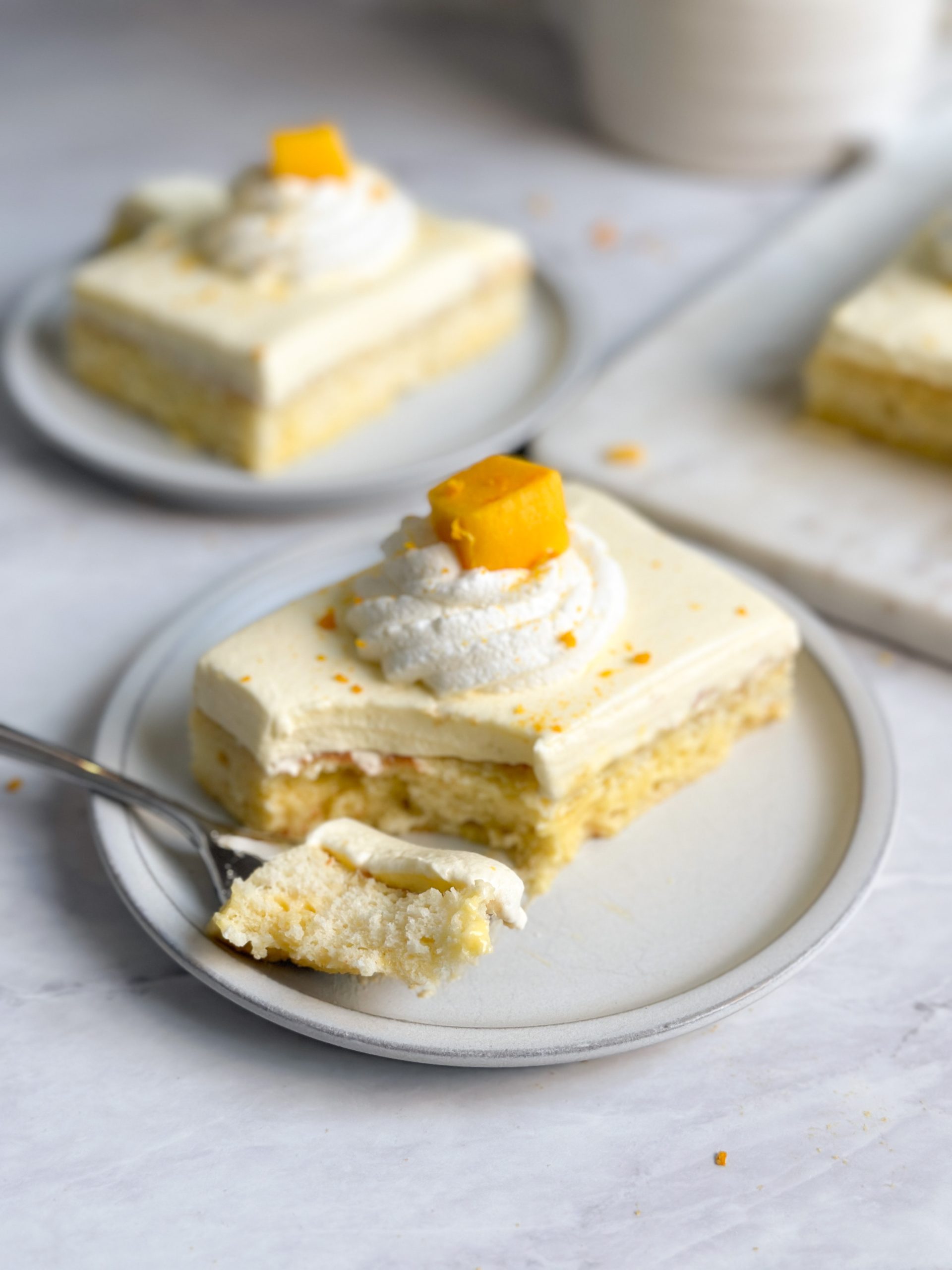 a slice of mango lassi tres leches cake on a plate with a moist interior, thick layer of mango lassi whipped cream, and whipped cream and mango decoration