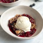berry crumble in a bowl with a crispy oat crumble and bubbling berries underneath. topped with a scoop of vanilla icecream