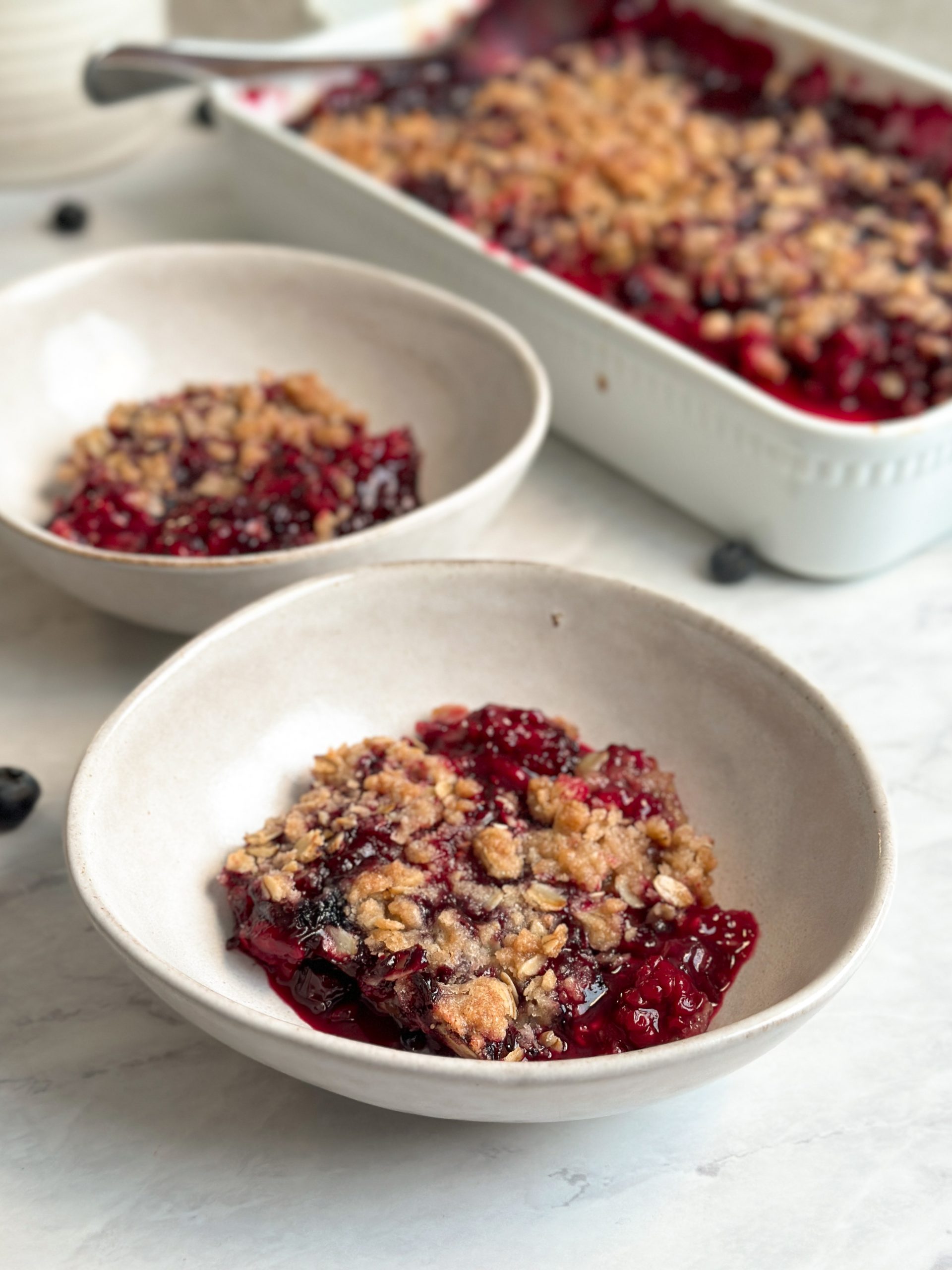 berry crumble in a bowl with a crispy oat crumble and bubbling berries underneath. Another bowl and dish seen in the background