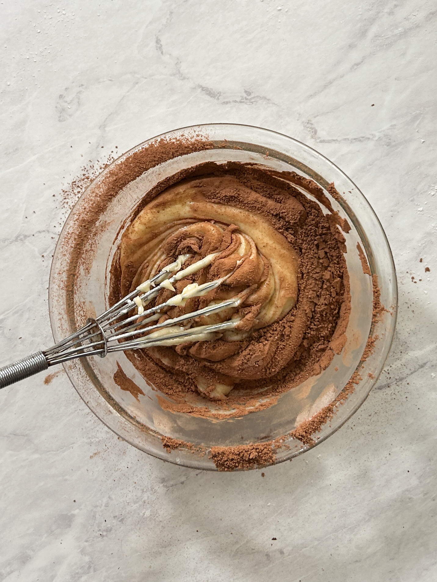 hand whisk being used to fold cocoa powder into cake batter in a glass bowl