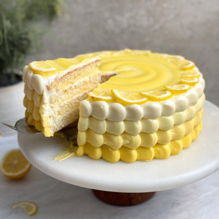 lemon cake on a marble cake stand with a spatula pulling out 1 slice revealing 4 layers o cake, frosting and lemon curd