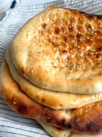 pile of 4 roghni naans on a tea towel. Naans have a beautiful golden color and sesame seeds on them