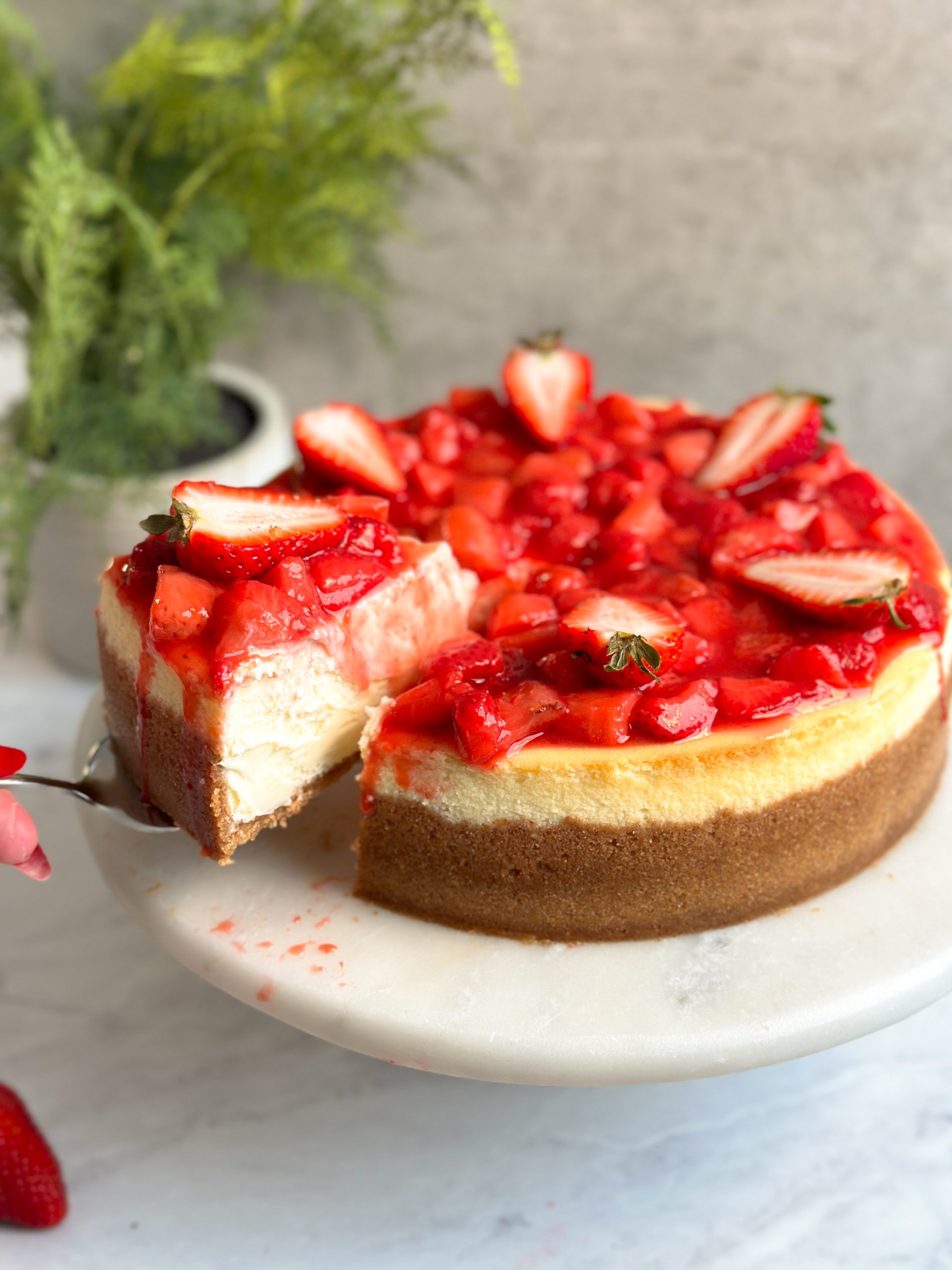 strawberry cheesecake on a serving stand with strawberry compote and fresh strawberries on top. A slice being taken out revealing creamy interior