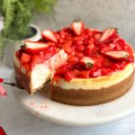 strawberry cheesecake on a serving stand with strawberry compote and fresh strawberries on top. A slice being taken out revealing creamy interior
