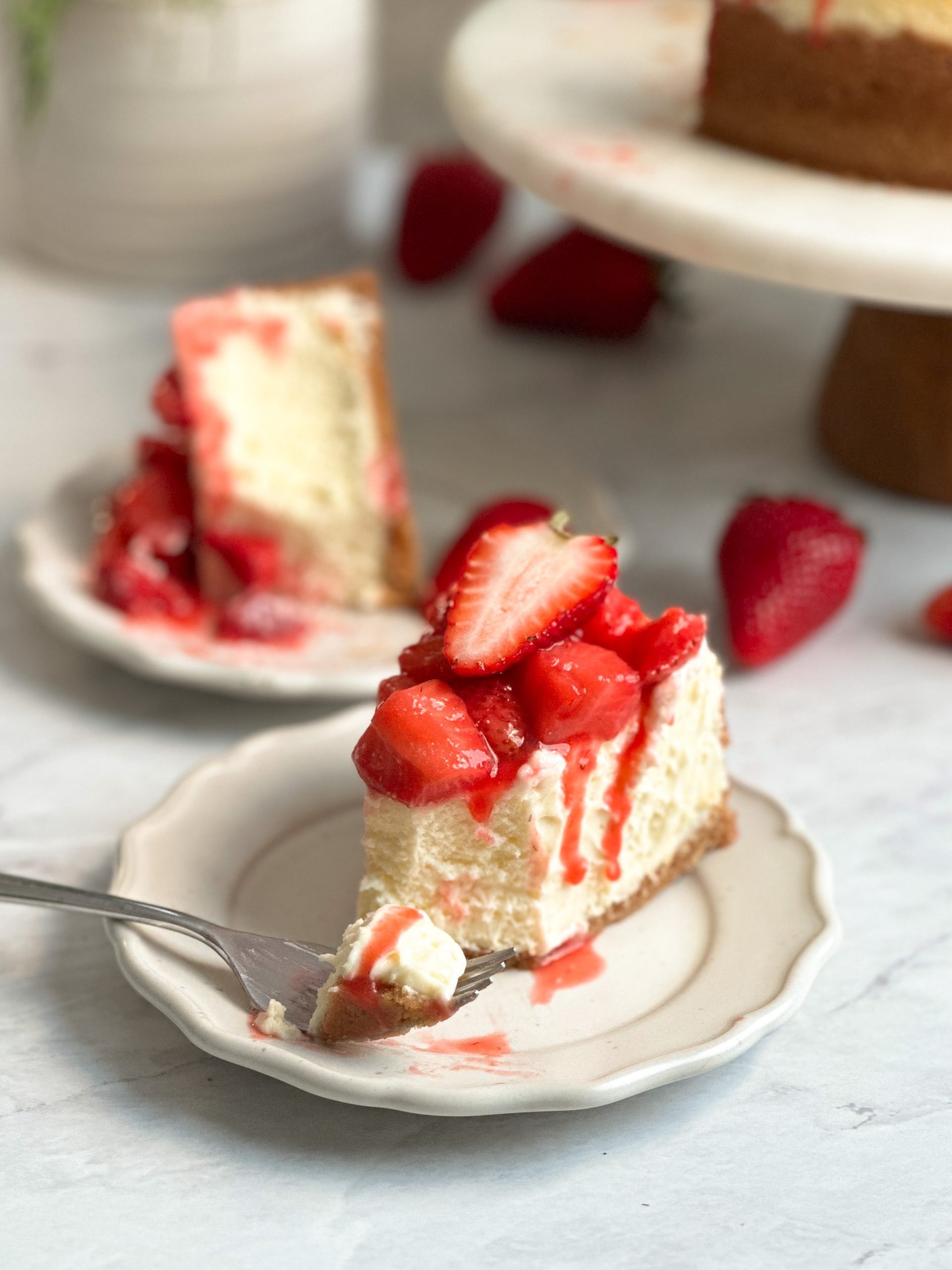 slice of cheesecake on a small plate with a fork taking a bite. cheesecake has a very creamy texture and is covered with a saucy strawberry compote