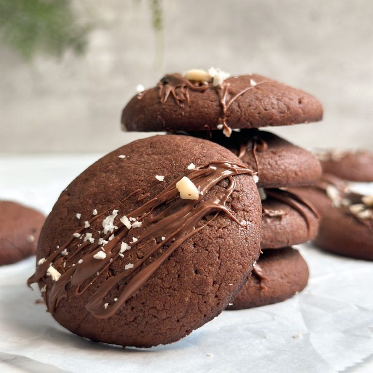 stack of 4 small round chewy nutella cookies, with another cookie perched on the edge, decorated with nutella and hazelnuts