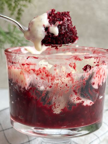 a glass mug from the side with a deep red red velvet cake inside with cream cheese frosting, spoon is pulling out a bite revealing moist texture inside