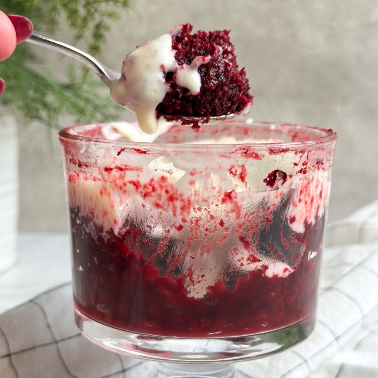 a glass mug from the side with a deep red red velvet cake inside with cream cheese frosting, spoon is pulling out a bite revealing moist texture inside