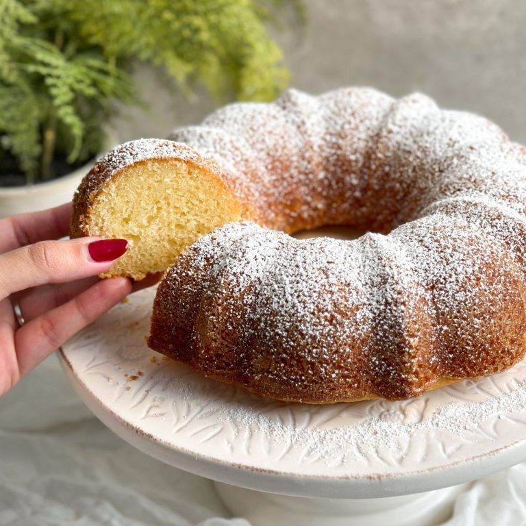 classic vanilla bundt cake on marble serving stand dusted with powdered sugar; hand pulling out 1 slice revealing soft texture inside