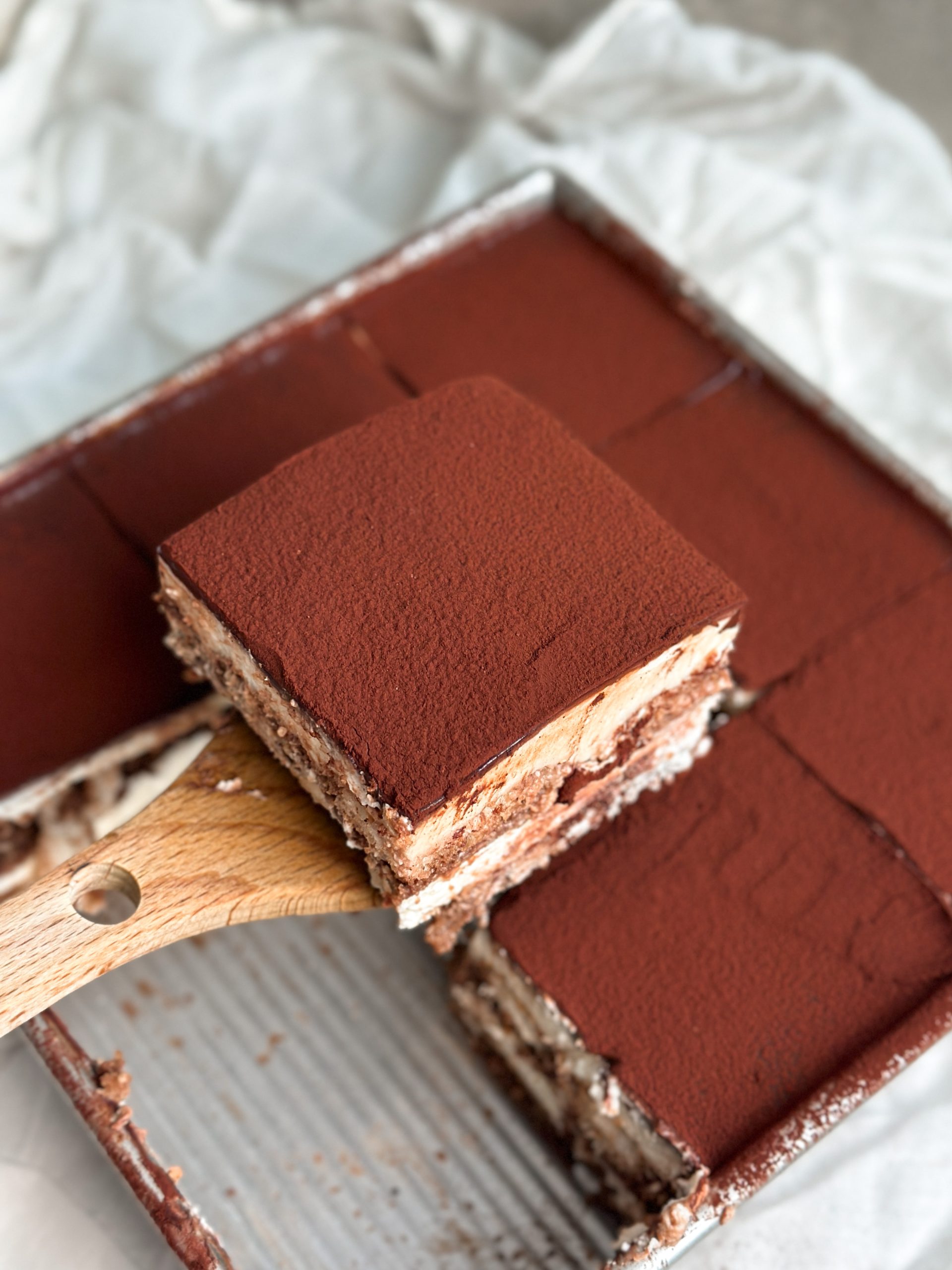 chocolate tiramisu in a 9x9" baking dish cut into slices, spatula pulling out one slice revealing soft layers of cream, ganache and ladyfingers
