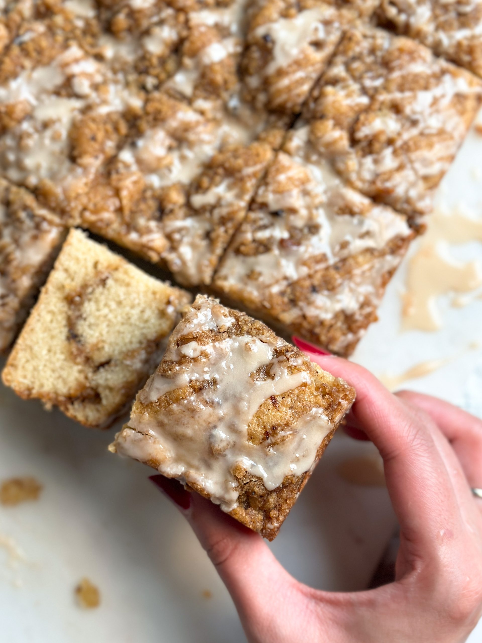 slices of Coffee cake with coffee streusel topping and coffee icing on a marble serving board, with one slice turned sideways to reveal moist and soft interior and more streusel inside; hand removing 1 slice