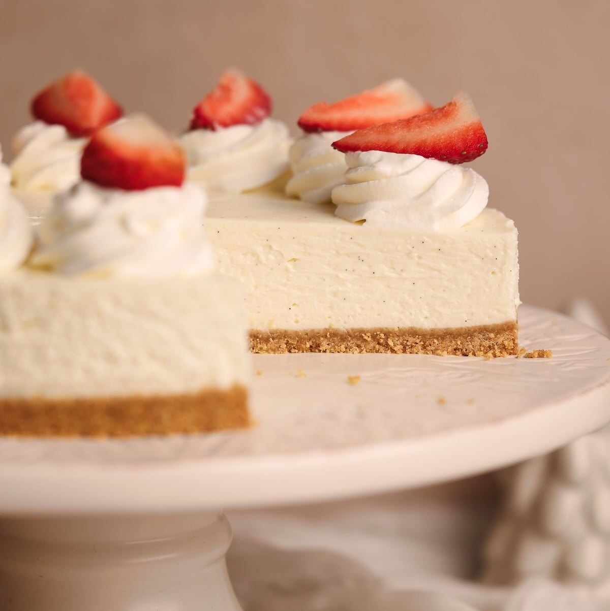 A close-up of the best no-bake cheesecake on a white cake stand, topped with swirls of whipped cream and sliced strawberries. A slice has been removed, revealing the creamy filling and graham cracker crust.