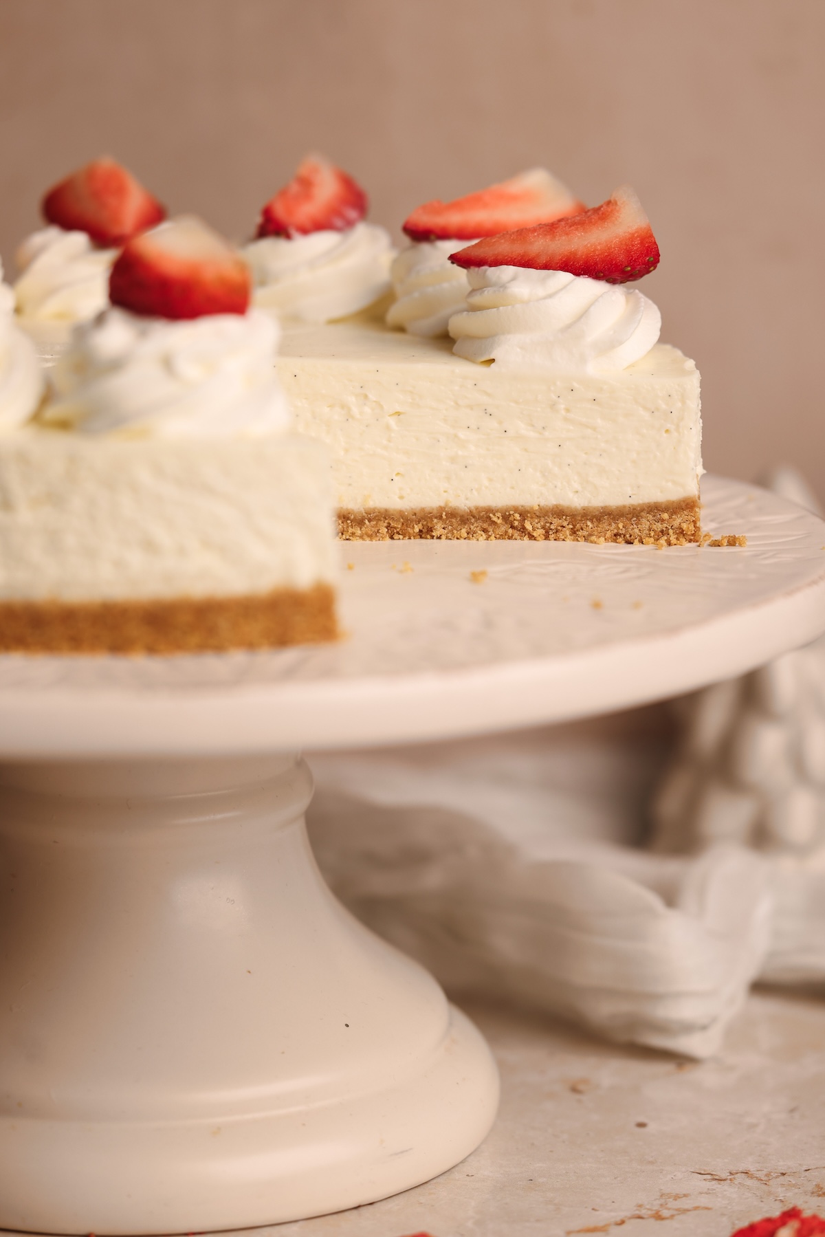 A best no-bake cheesecake with a graham cracker crust, topped with whipped cream and sliced strawberries, sits on a white cake stand. One slice has been removed.