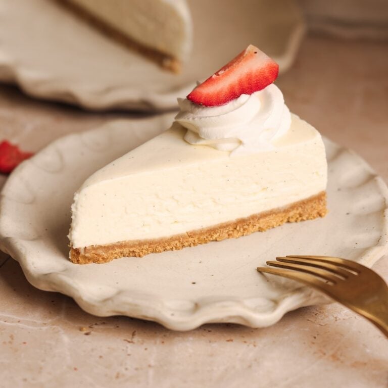 A slice of no-bake cheesecake with a graham cracker crust, topped with a swirl of whipped cream and a strawberry slice, served on a ceramic plate with a gold fork beside it.