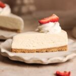 A slice of creamy, best no-bake cheesecake with a graham cracker crust, topped with a swirl of whipped cream and a sliced strawberry, sits on a white plate.