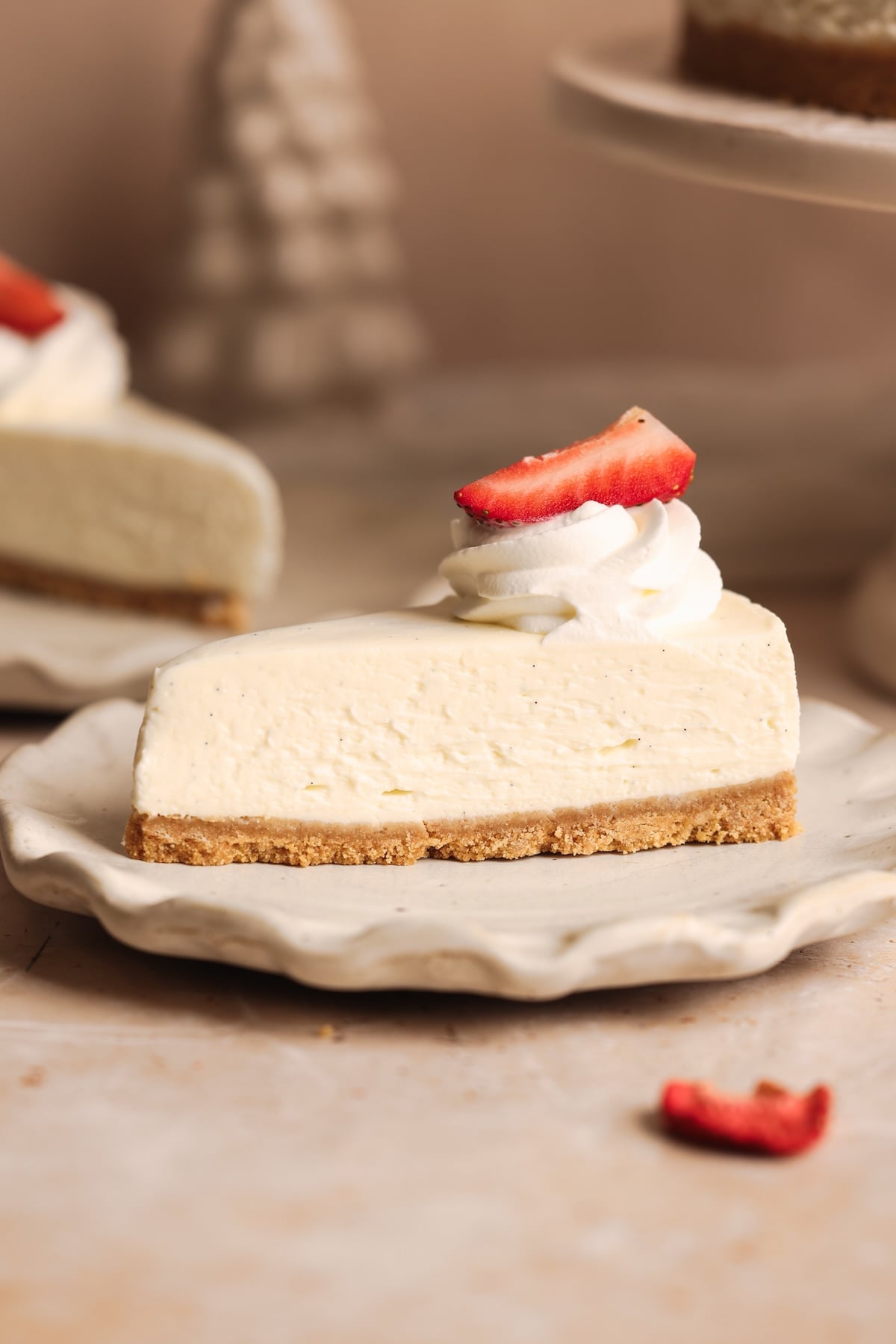 A slice of the best no-bake cheesecake with a graham cracker crust, topped with a swirl of whipped cream and a strawberry slice, sits on a ceramic plate. Another cheesecake is partially visible in the background.