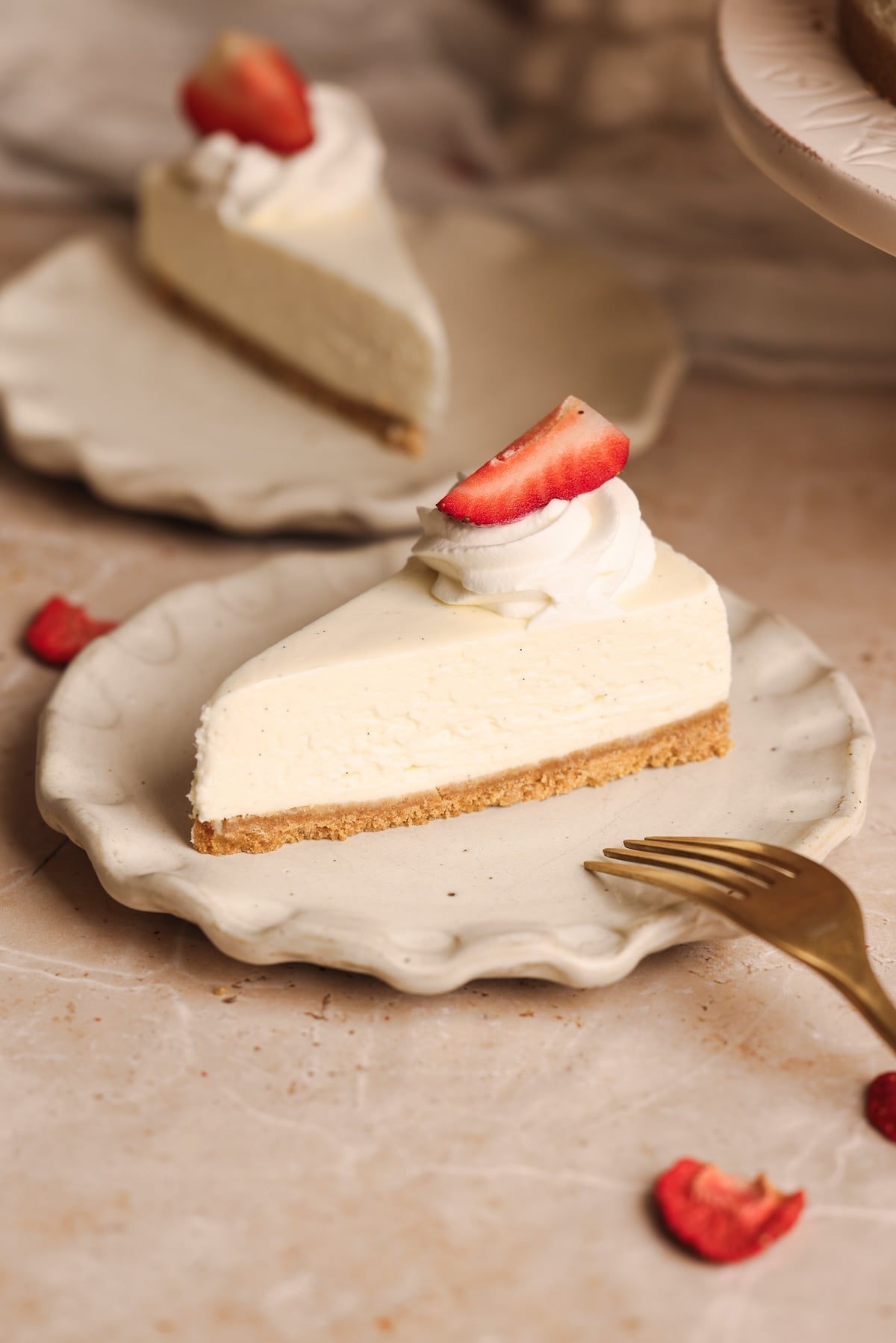 A slice of the best no-bake cheesecake with a graham cracker crust, topped with a swirl of whipped cream and a sliced strawberry, sits on a decorative plate next to a gold fork. Another tempting slice is blurred in the background.