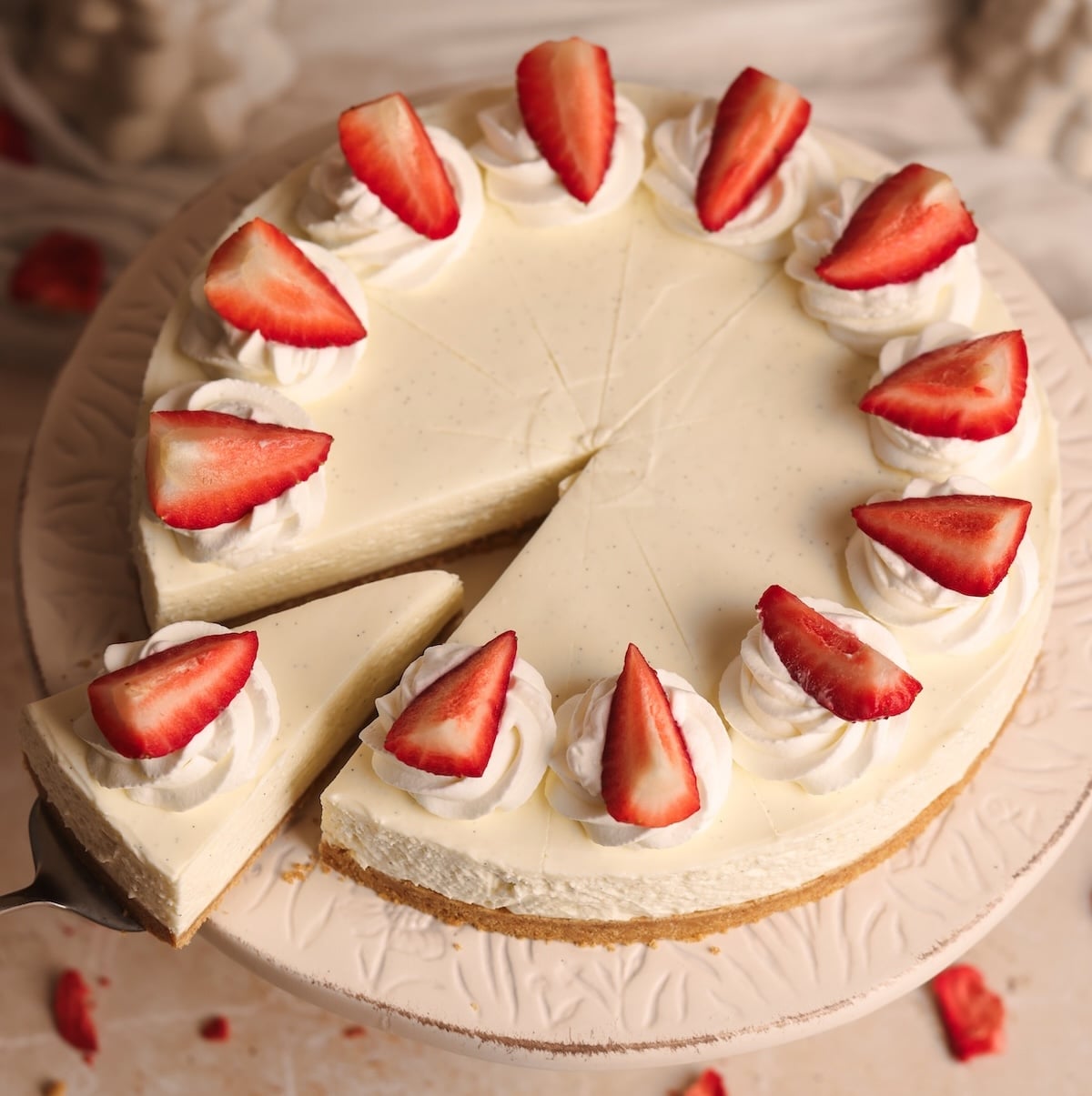 A creamy no-bake cheesecake topped with whipped cream and halved strawberries, with one slice being served on a pie server. The cake sits on a decorative white plate.