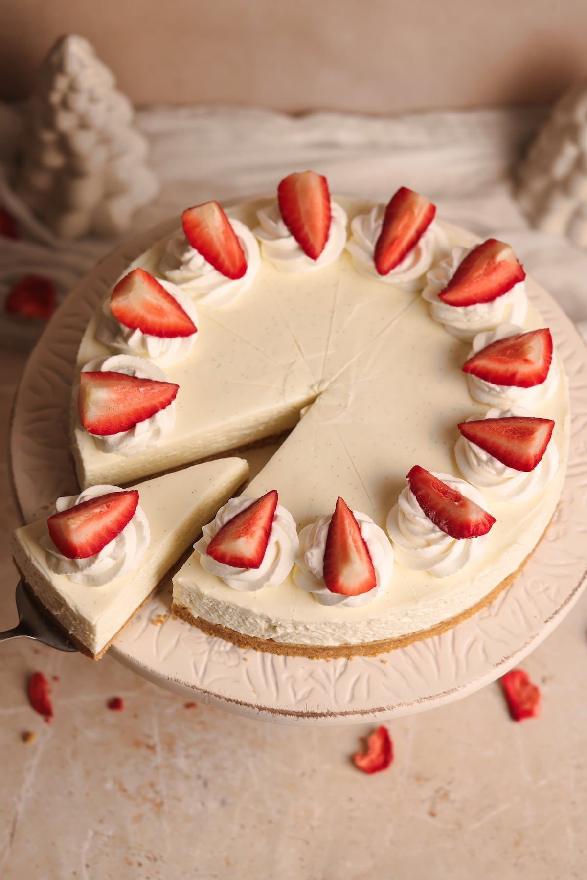A best no-bake cheesecake topped with whipped cream and halved strawberries sits on a decorative cake stand. One slice is being lifted from the cake, with small pieces of strawberry scattered nearby.
