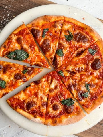 overhead image of pizza margherita on a round marble serving board. pizza has a crispy crust, melted cheese and basil leaves on it. one slice has been pulled out