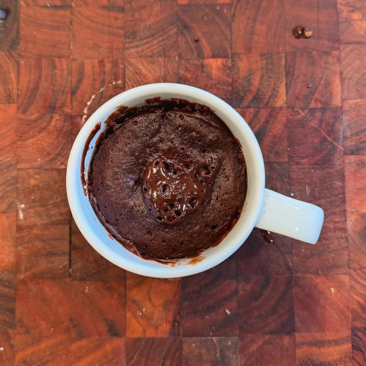 A white mug filled with a cooked chocolate mug cake sits on a wooden patterned surface. The cake has a slightly moist, bubbly center, and some chocolate smudges are visible on the mug's rim.