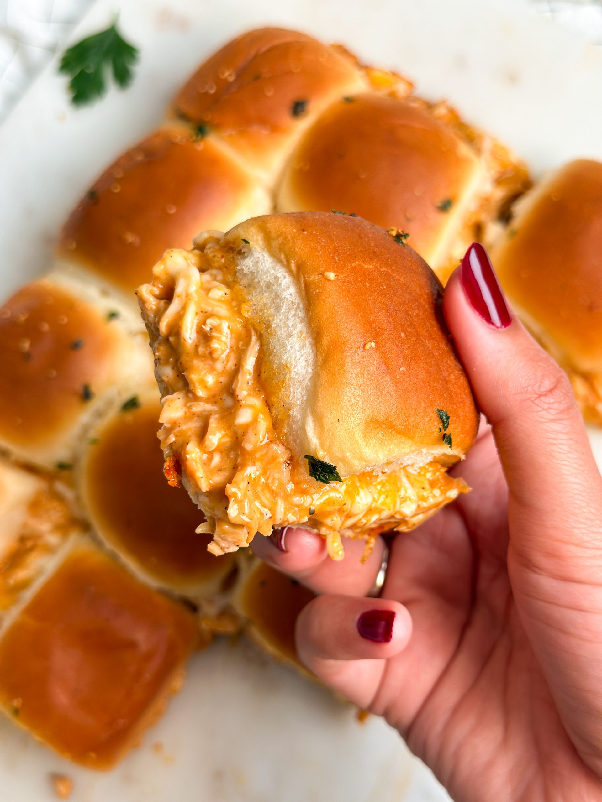 hand holding a bbq chicken slider sideways to show the cheesy filling with more rolls in the background