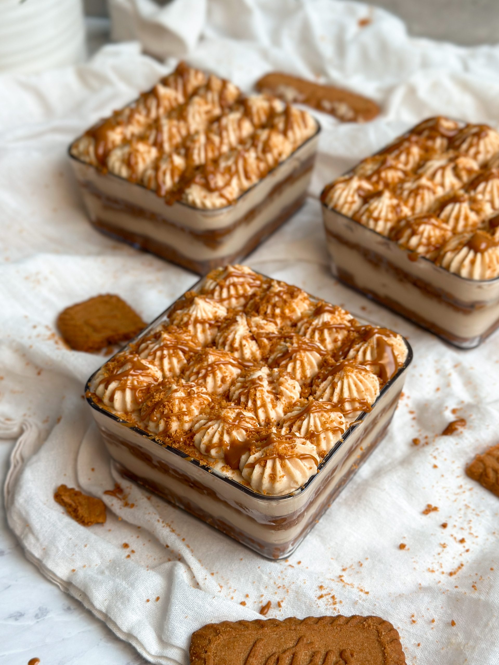3 square dessert cups with biscoff tiramisu . layers of biscoff cream and cookies seen from the side. decorated with piping and crushed biscoff cookies and drizzled cookie butter. one cup is in focus in the front placed facing the camera