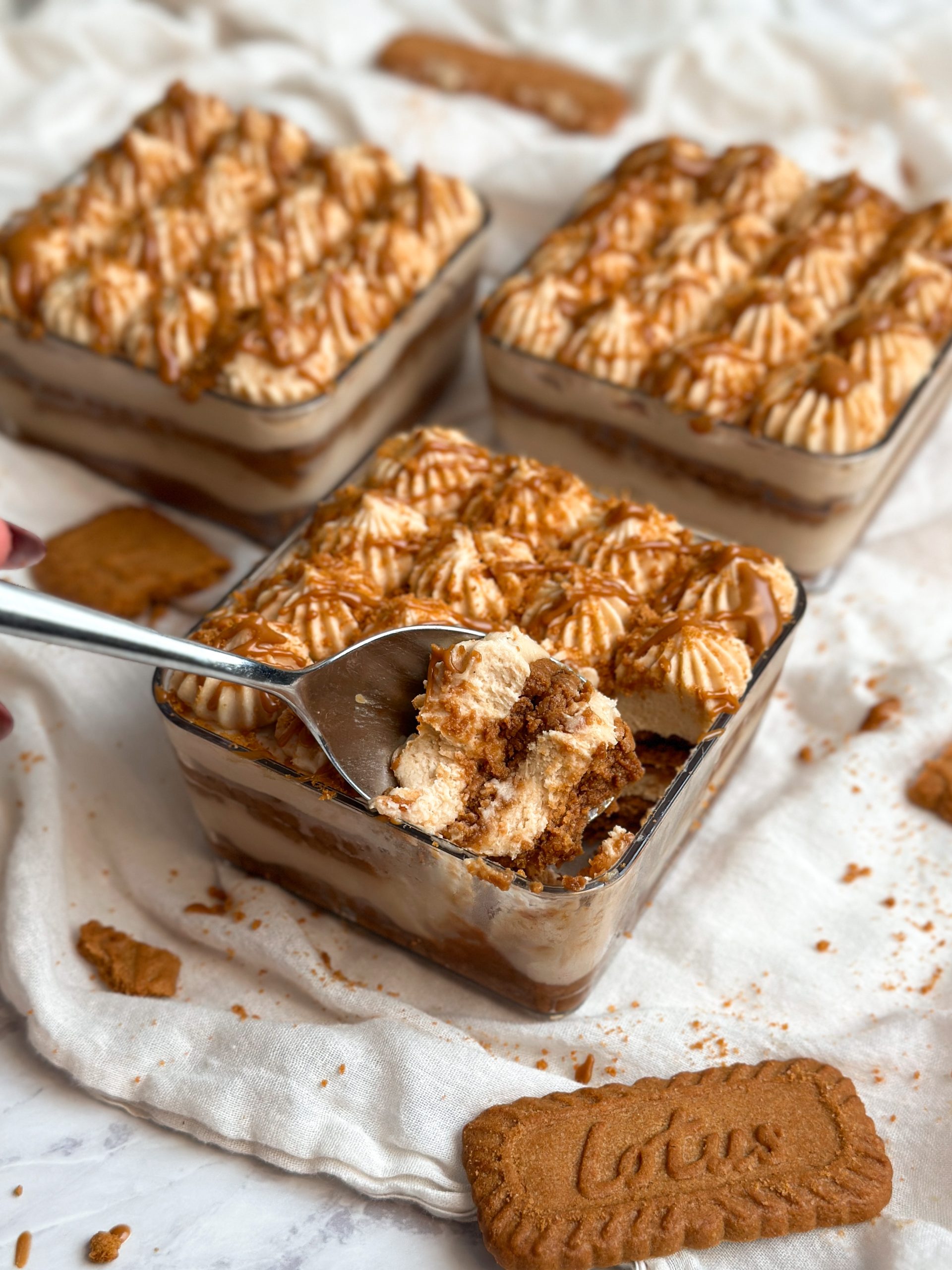 3 square dessert cups with biscoff tiramisu . layers of biscoff cream and cookies seen from the side. decorated with piping and crushed biscoff cookies and drizzled cookie butter. one cup is in focus in the front with a spoon taking out a bite from it