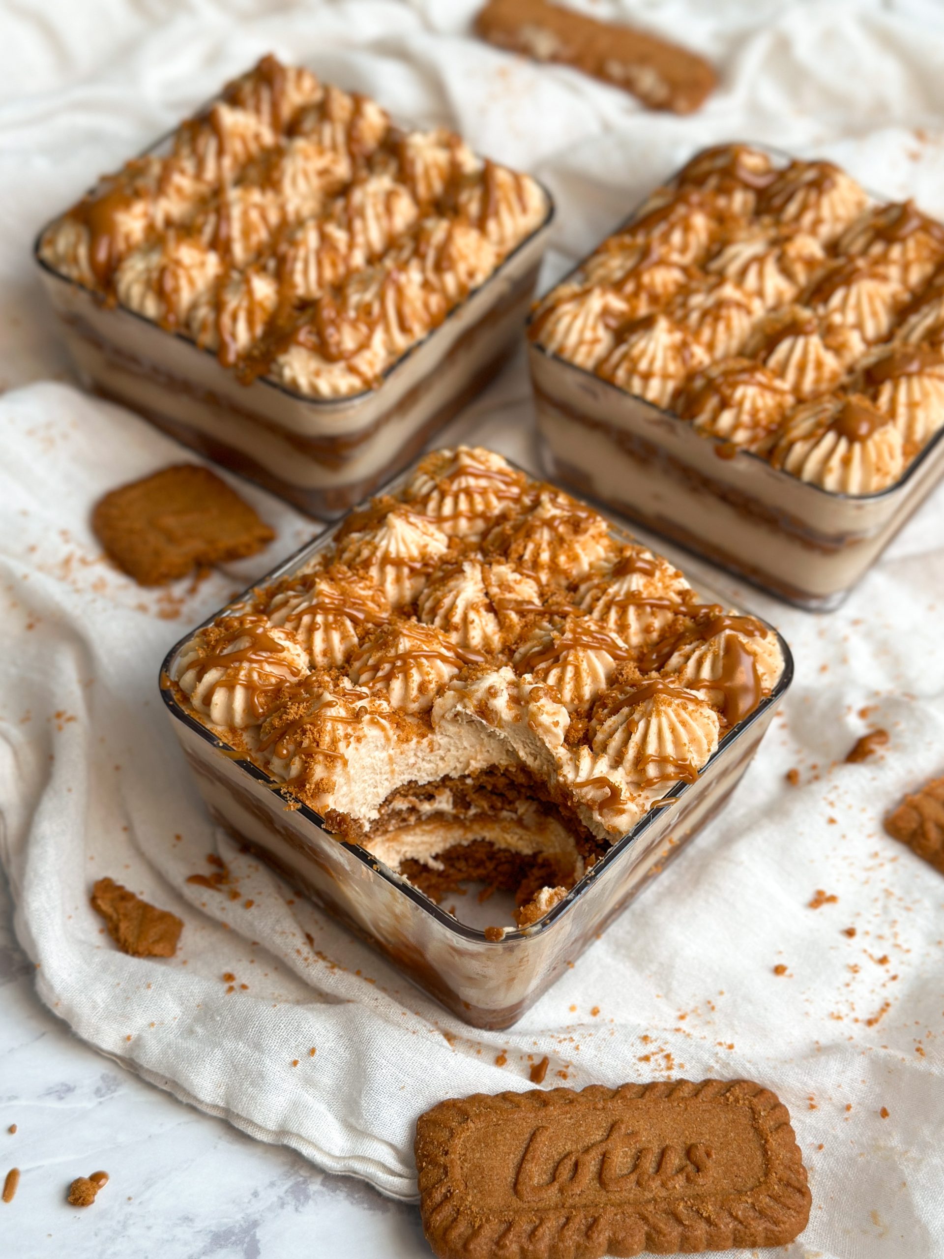 3 square dessert cups with biscoff tiramisu . layers of biscoff cream and cookies seen from the side. decorated with piping and crushed biscoff cookies and drizzled cookie butter. one cup is in focus in the front with a bite taken out revealing the creamy layers inside