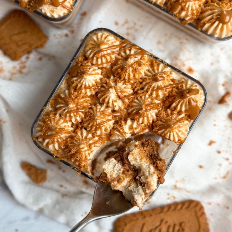 biscoff tiramisu dessert from the top in a dessert cup with pretty piping of biscoff cream, crushed biscoff cookies, and drizzled cookie butter. a spoon is taking out a bite revealing layers of cream and cookies inside
