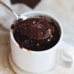 A spoon lifts a portion of gooey chocolate mug cake from a white mug, showing a moist, rich texture. The mug sits on a white saucer, with melted chocolate visible on the cake and rim.