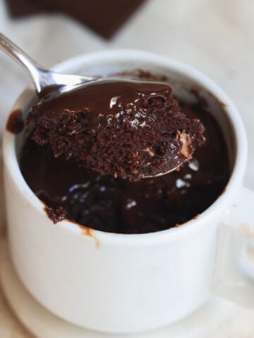 A spoon lifts a portion of gooey chocolate mug cake from a white mug, showing a moist, rich texture. The mug sits on a white saucer, with melted chocolate visible on the cake and rim.