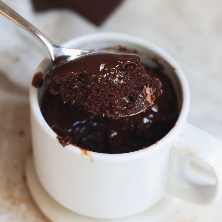 A spoon lifts a portion of gooey chocolate mug cake from a white mug, showing a moist, rich texture. The mug sits on a white saucer, with melted chocolate visible on the cake and rim.