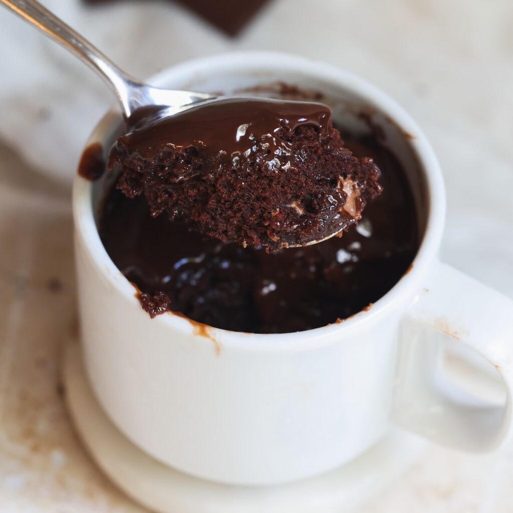 A spoon holds a bite of rich, gooey chocolate mug cake above a white mug filled with more cake. This chocolate mug cake looks irresistibly moist and chocolatey, with some melted chocolate on top.