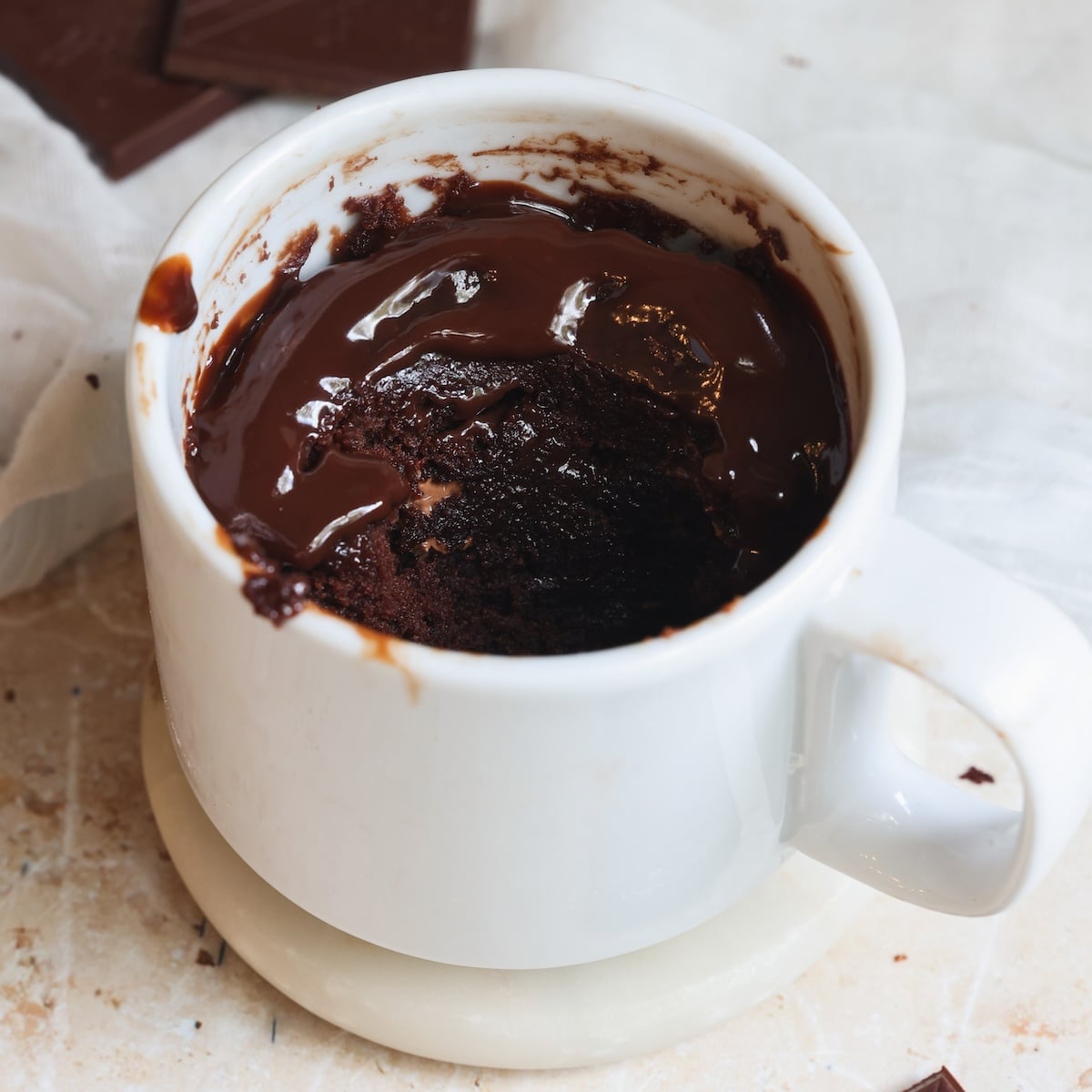 A white mug filled with a rich, gooey chocolate mug cake, partially eaten, sits on a white coaster. Pieces of chocolate and a white cloth are visible in the background.